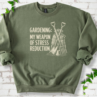 Military Green sweatshirt flat lay with cream "Gardening: My Weapon of Stress Reduction" lettering beside a crossed spade and fork, burlap sack and a small flowerpot, edged by green leaves.