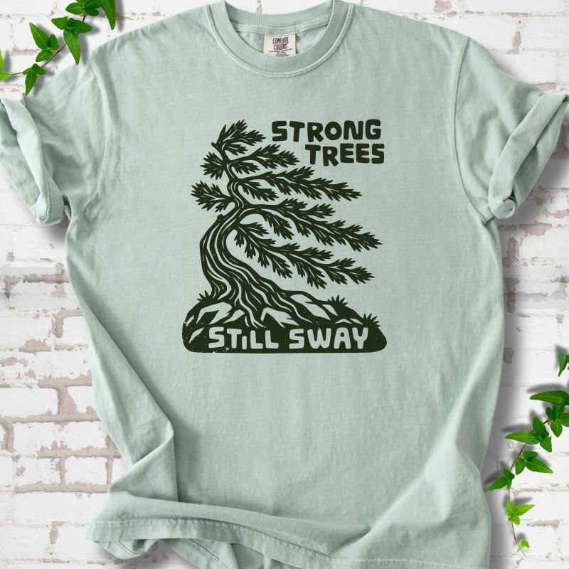 Front flat lay of a Bay short sleeve T-shirt with a wind-bent tree, exposed roots and Strong Trees Still Sway lettering, on white brick with green leafy sprigs.