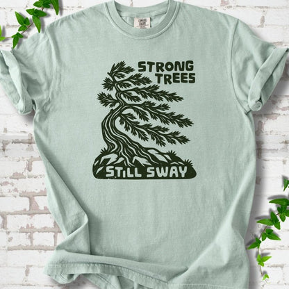 Front flat lay of a Bay short sleeve T-shirt with a wind-bent tree, exposed roots and Strong Trees Still Sway lettering, on white brick with green leafy sprigs.