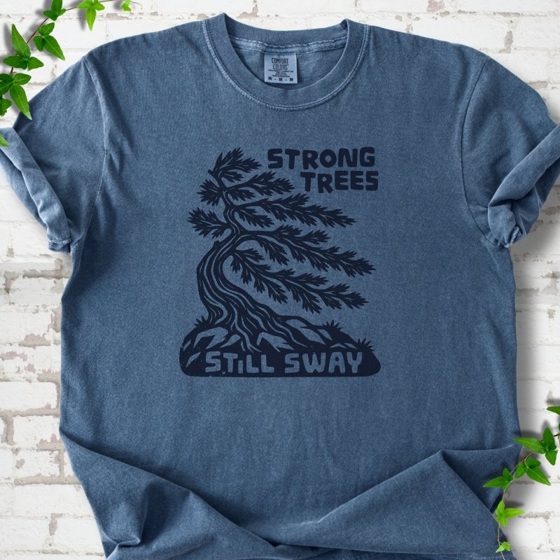 Front flat lay of a Denim short sleeve T-shirt with a wind-bent tree, exposed roots and Strong Trees Still Sway lettering, on white brick with green leafy sprigs.