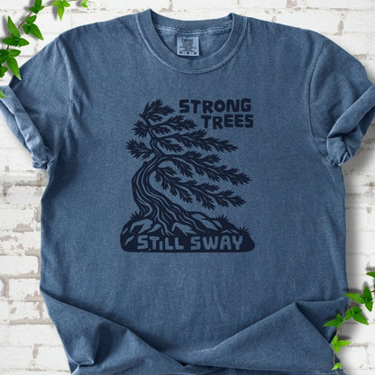 Front flat lay of a Denim short sleeve T-shirt with a wind-bent tree, exposed roots and Strong Trees Still Sway lettering, on white brick with green leafy sprigs.