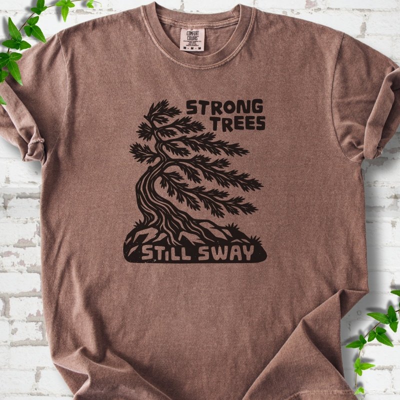 Front flat lay of a Espresso short sleeve T-shirt with a wind-bent tree, exposed roots and Strong Trees Still Sway lettering, on white brick with green leafy sprigs.