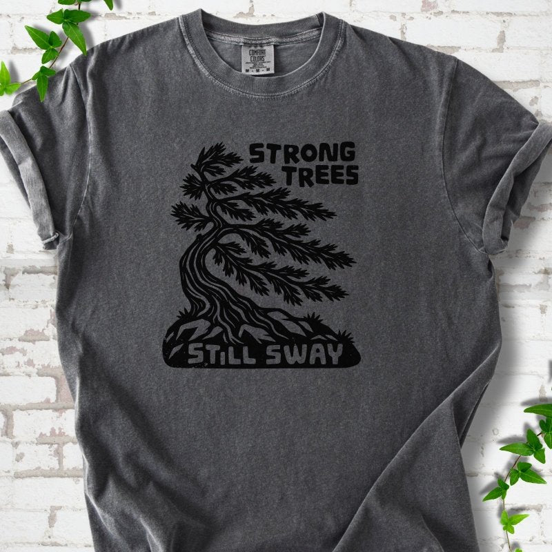 Front flat lay of a Pepper short sleeve T-shirt with a wind-bent tree, exposed roots and Strong Trees Still Sway lettering, on white brick with green leafy sprigs.