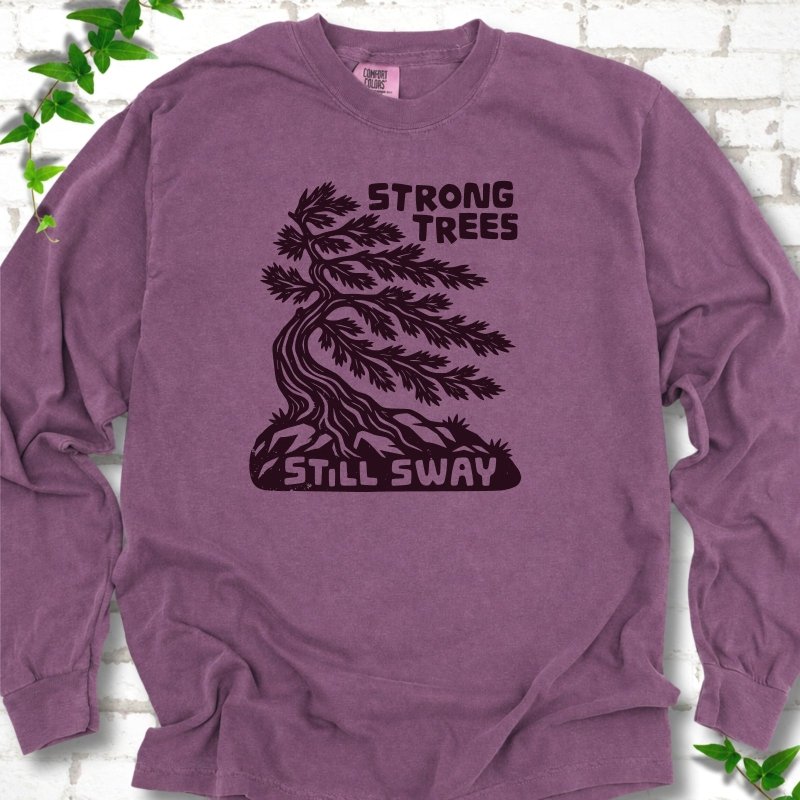 Front flat lay of a Berry long sleeve T-shirt with a wind-bent tree, exposed roots and Strong Trees Still Sway lettering, on white brick with green leafy sprigs.