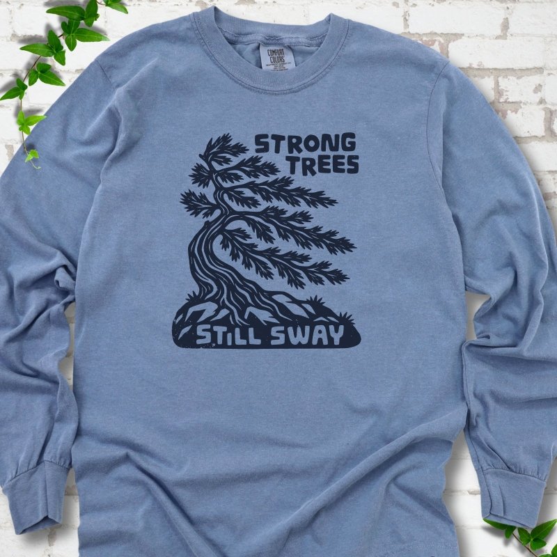 Front flat lay of a Blue Jean long sleeve T-shirt with a wind-bent tree, exposed roots and Strong Trees Still Sway lettering, on white brick with green leafy sprigs.