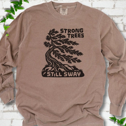 Front flat lay of a Espresso long sleeve T-shirt with a wind-bent tree, exposed roots and Strong Trees Still Sway lettering, on white brick with green leafy sprigs.