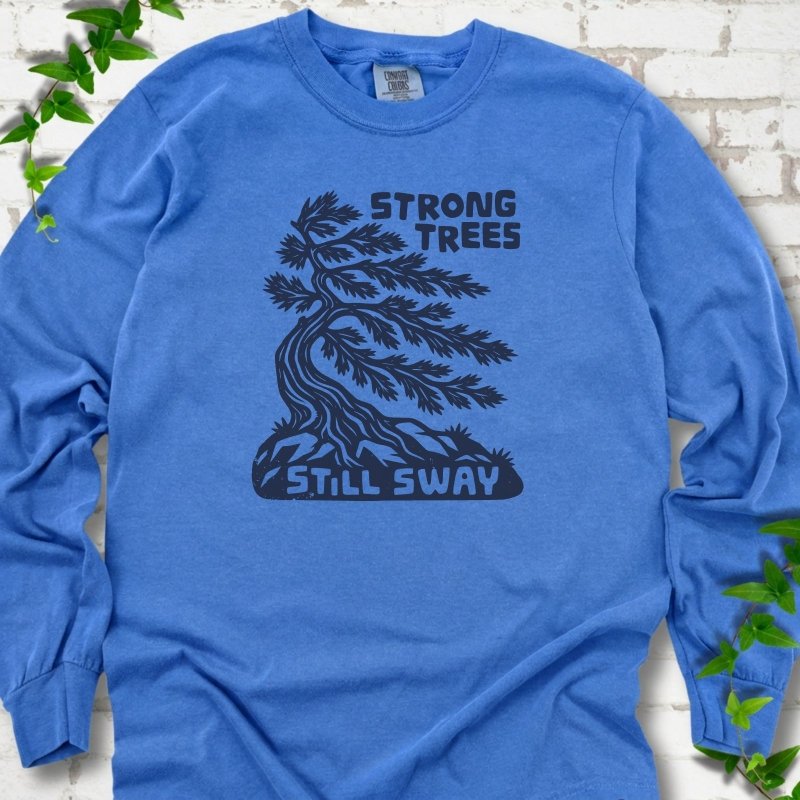 Front flat lay of a Flo Blue long sleeve T-shirt with a wind-bent tree, exposed roots and Strong Trees Still Sway lettering, on white brick with green leafy sprigs.