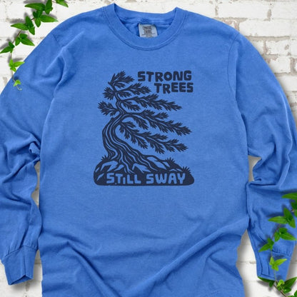 Front flat lay of a Flo Blue long sleeve T-shirt with a wind-bent tree, exposed roots and Strong Trees Still Sway lettering, on white brick with green leafy sprigs.