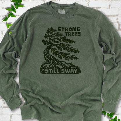 Front flat lay of a Hemp long sleeve T-shirt with a wind-bent tree, exposed roots and Strong Trees Still Sway lettering, on white brick with green leafy sprigs.