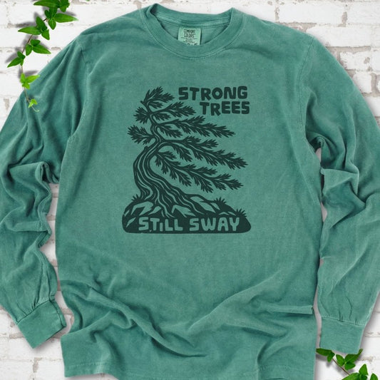 Front flat lay of a Light Green long sleeve T-shirt with a wind-bent tree, exposed roots and Strong Trees Still Sway lettering, on white brick with green leafy sprigs.
