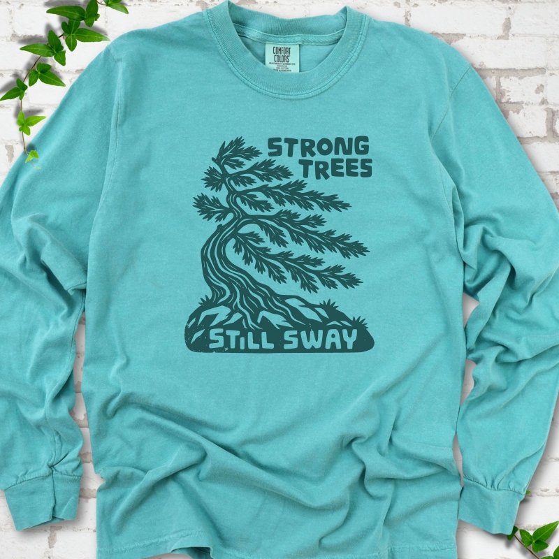 Front flat lay of a Seafoam long sleeve T-shirt with a wind-bent tree, exposed roots and Strong Trees Still Sway lettering, on white brick with green leafy sprigs.