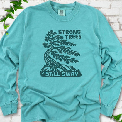Front flat lay of a Seafoam long sleeve T-shirt with a wind-bent tree, exposed roots and Strong Trees Still Sway lettering, on white brick with green leafy sprigs.