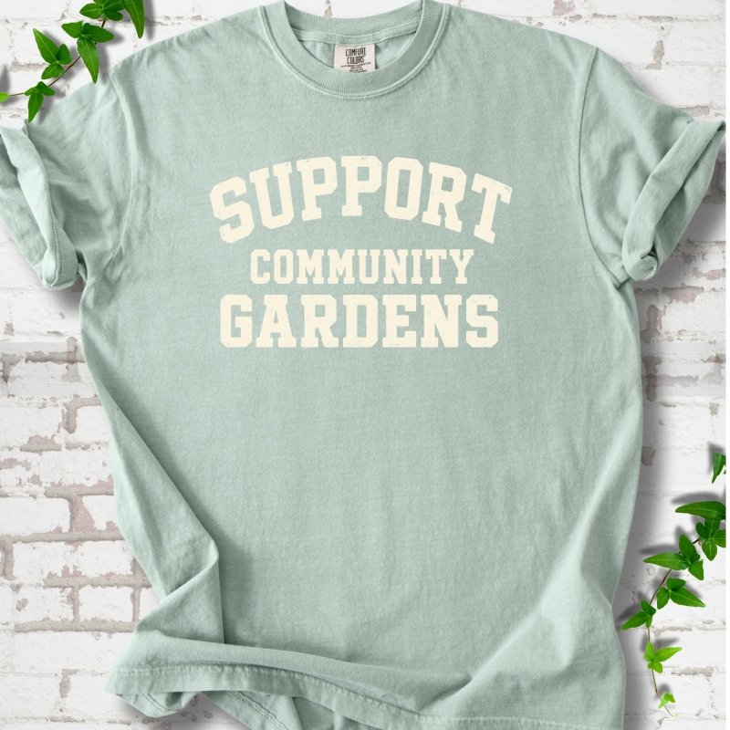 Bay light green short-sleeve T-shirt with Support Community Gardens in cream collegiate block letters across the chest, flat lay on white brick with ivy