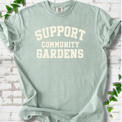 Bay light green short-sleeve T-shirt with Support Community Gardens in cream collegiate block letters across the chest, flat lay on white brick with ivy