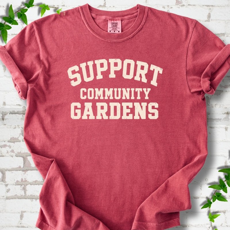 Crimson red short-sleeve T-shirt with Support Community Gardens in cream collegiate block letters across the chest, flat lay on white brick with ivy