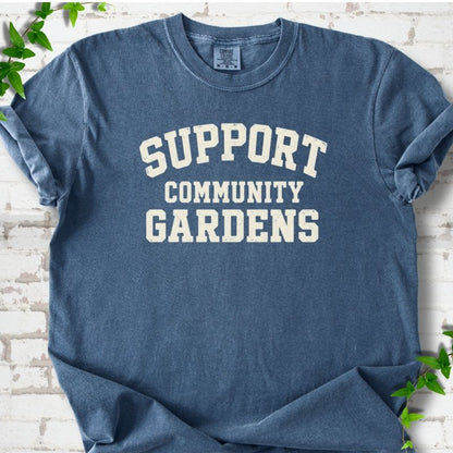 Denim blue short-sleeve T-shirt with Support Community Gardens in cream collegiate block letters across the chest, flat lay on white brick with ivy