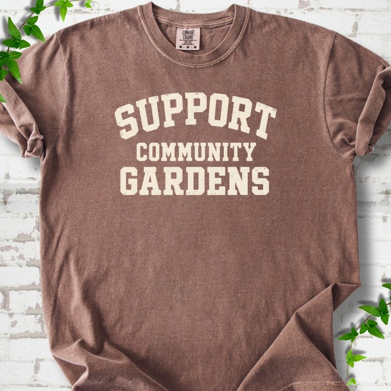 Espresso brown short-sleeve T-shirt with Support Community Gardens in cream collegiate block letters across the chest, flat lay on white brick with ivy