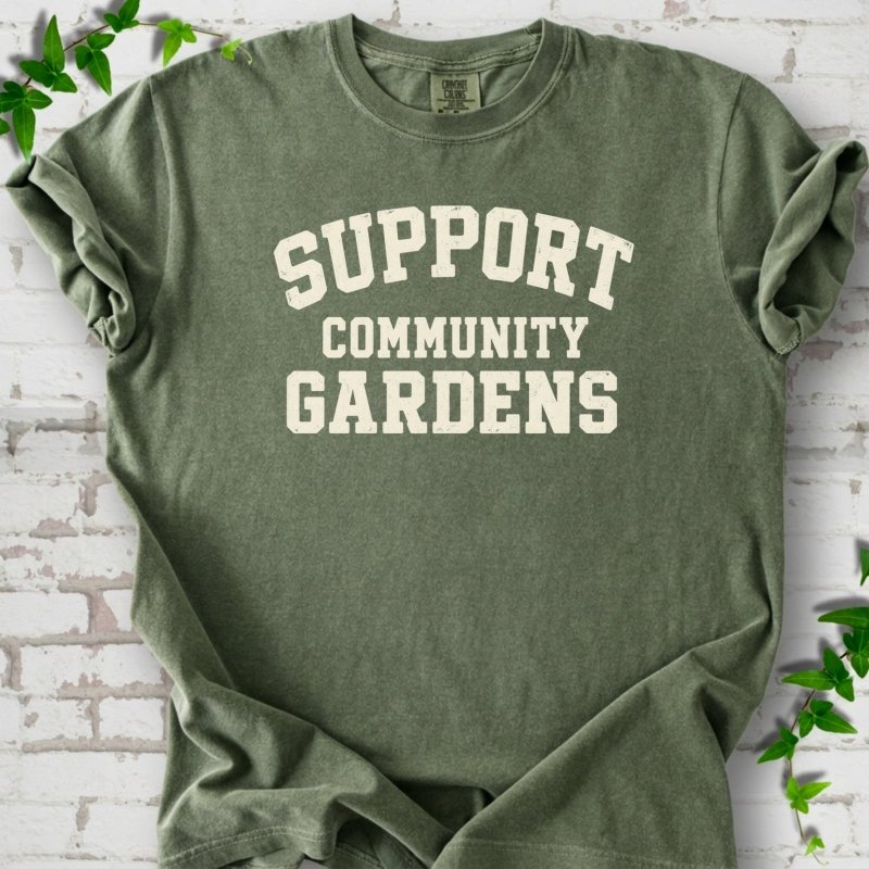 Moss green short-sleeve T-shirt with Support Community Gardens in cream collegiate block letters across the chest, flat lay on white brick with ivy