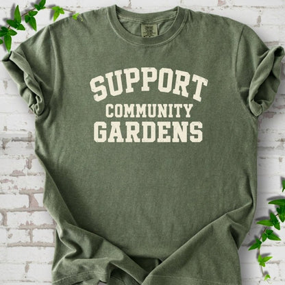 Moss green short-sleeve T-shirt with Support Community Gardens in cream collegiate block letters across the chest, flat lay on white brick with ivy