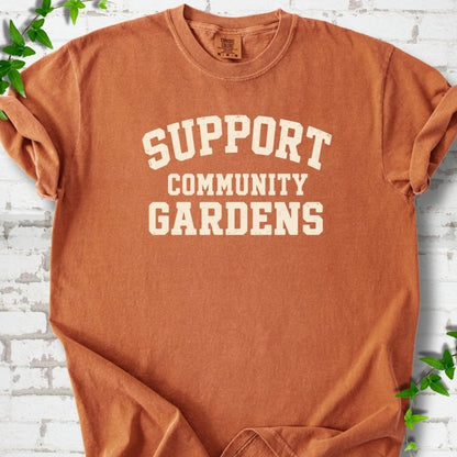 Yam burnt orange short-sleeve T-shirt with Support Community Gardens in cream collegiate block letters across the chest, flat lay on white brick with ivy