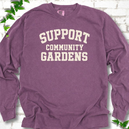 Berry plum long sleeve T-shirt with Support Community Gardens in cream collegiate block letters across the chest, flat lay on white brick with ivy