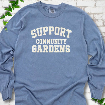 Blue Jean light blue long sleeve T-shirt with Support Community Gardens in cream collegiate block letters across the chest, flat lay on white brick with ivy
