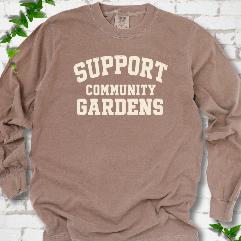 Espresso brown long sleeve T-shirt with Support Community Gardens in cream collegiate block letters across the chest, flat lay on white brick with ivy
