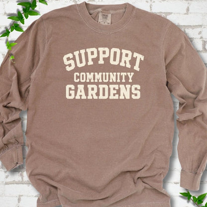 Espresso brown long sleeve T-shirt with Support Community Gardens in cream collegiate block letters across the chest, flat lay on white brick with ivy