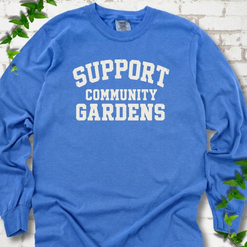 Flo Blue bright blue long sleeve T-shirt with Support Community Gardens in cream collegiate block letters across the chest, flat lay on white brick with ivy