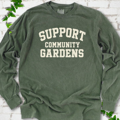 Hemp green long sleeve T-shirt with Support Community Gardens in cream collegiate block letters across the chest, flat lay on white brick with ivy