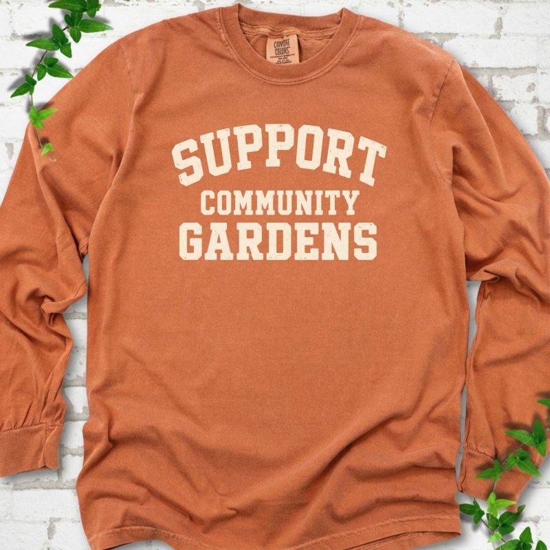 Yam burnt orange long sleeve T-shirt with Support Community Gardens in cream collegiate block letters across the chest, flat lay on white brick with ivy