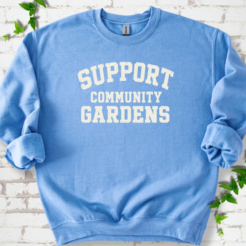 Carolina blue crewneck sweatshirt with Support Community Gardens in cream collegiate block letters across the chest, flat lay on white brick with ivy