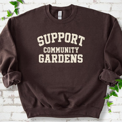Dark chocolate brown crewneck sweatshirt with Support Community Gardens in cream collegiate block letters across the chest, flat lay on white brick with ivy