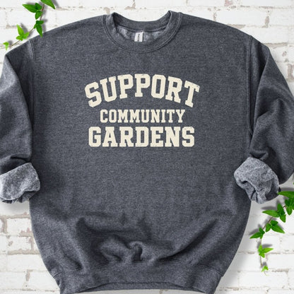 Dark heather charcoal gray crewneck sweatshirt with Support Community Gardens in cream collegiate block letters across the chest, flat lay on white brick with ivy
