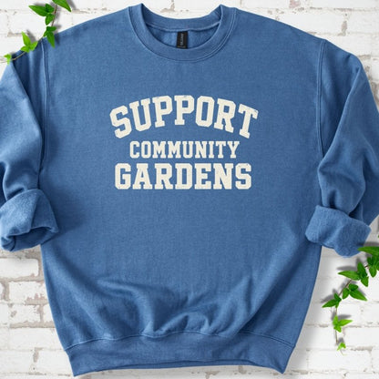 Indigo blue crewneck sweatshirt with Support Community Gardens in cream collegiate block letters across the chest, flat lay on white brick with ivy