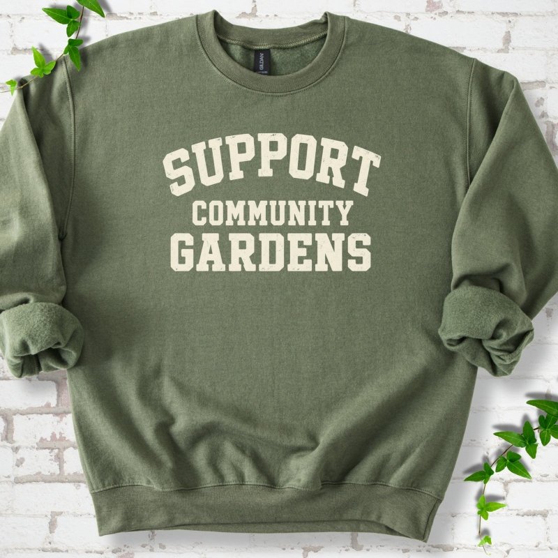 Military green crewneck sweatshirt with Support Community Gardens in cream collegiate block letters across the chest, flat lay on white brick with ivy