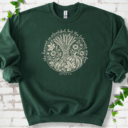 Forest Green sweatshirt flat lay with cream wheat, pumpkins and leaves in a harvest circle, the Matthew 9:37 verse around it and its reference below.