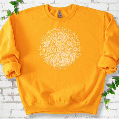 Gold sweatshirt flat lay with cream wheat, pumpkins and leaves in a harvest circle, the Matthew 9:37 verse around it and its reference below.