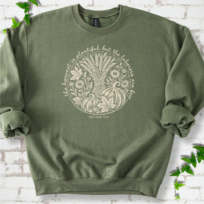 Military Green sweatshirt flat lay with cream wheat, pumpkins and leaves in a harvest circle, the Matthew 9:37 verse around it and its reference below.