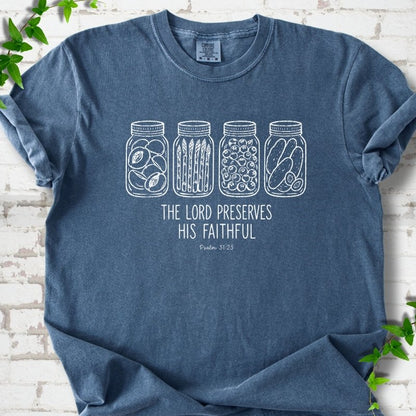 Denim T-shirt flat lay with white line-art illustrations of four mason jars filled with peaches, asparagus, blueberries and pickles above the words "The Lord Preserves His Faithful" and small "Psalm 31:23", on white brick with ivy sprigs.
