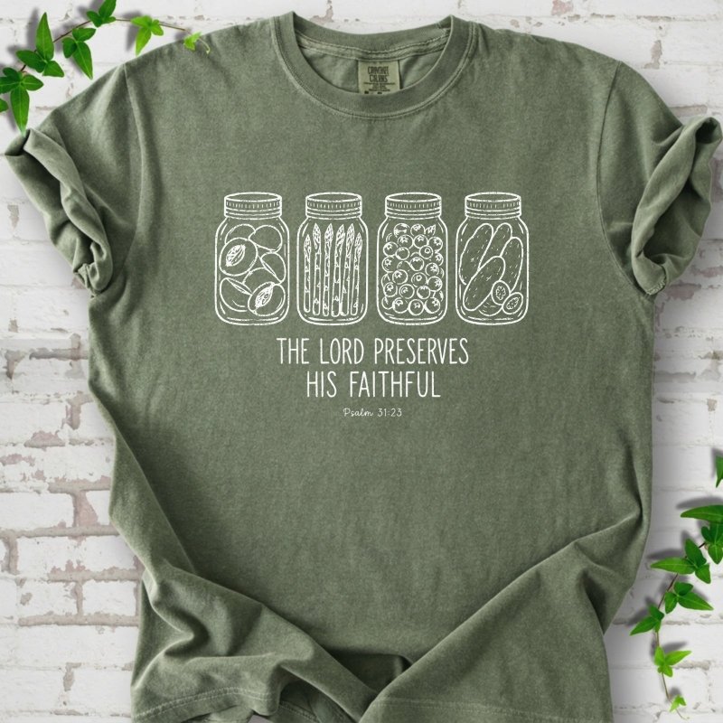 Moss T-shirt flat lay with white line-art illustrations of four mason jars filled with peaches, asparagus, blueberries and pickles above the words "The Lord Preserves His Faithful" and small "Psalm 31:23", on white brick with ivy sprigs.
