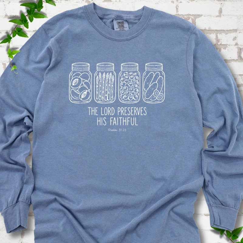Blue Jean long sleeve T-shirt flat lay with white line-art illustrations of four mason jars filled with peaches, asparagus, blueberries and pickles above the words "The Lord Preserves His Faithful" and small "Psalm 31:23", on white brick with ivy sprigs.