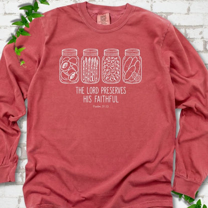 Crimson long sleeve T-shirt flat lay with white line-art illustrations of four mason jars filled with peaches, asparagus, blueberries and pickles above the words "The Lord Preserves His Faithful" and small "Psalm 31:23", on white brick with ivy sprigs.