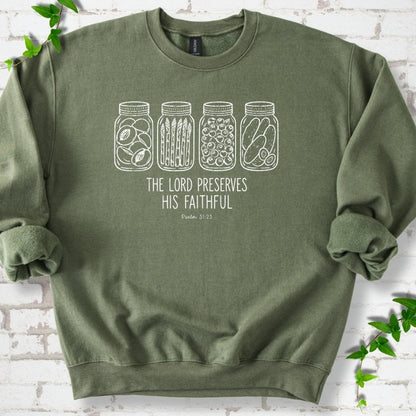 Military Green sweatshirt flat lay with white line-art illustrations of four mason jars filled with peaches, asparagus, blueberries and pickles above the words "The Lord Preserves His Faithful" and small "Psalm 31:23", on white brick with ivy sprigs.