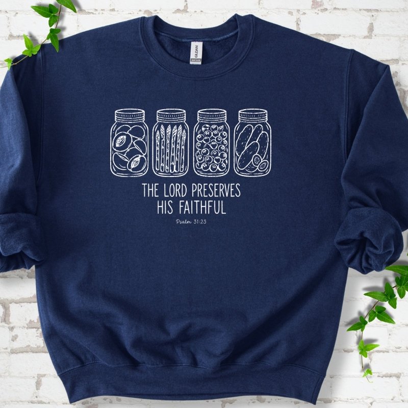 Navy sweatshirt flat lay with white line-art illustrations of four mason jars filled with peaches, asparagus, blueberries and pickles above the words "The Lord Preserves His Faithful" and small "Psalm 31:23", on white brick with ivy sprigs.
