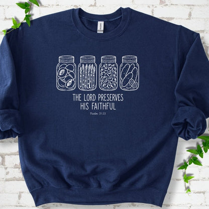 Navy sweatshirt flat lay with white line-art illustrations of four mason jars filled with peaches, asparagus, blueberries and pickles above the words "The Lord Preserves His Faithful" and small "Psalm 31:23", on white brick with ivy sprigs.