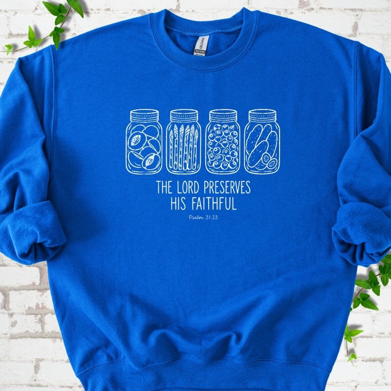 Royal sweatshirt flat lay with white line-art illustrations of four mason jars filled with peaches, asparagus, blueberries and pickles above the words "The Lord Preserves His Faithful" and small "Psalm 31:23", on white brick with ivy sprigs.