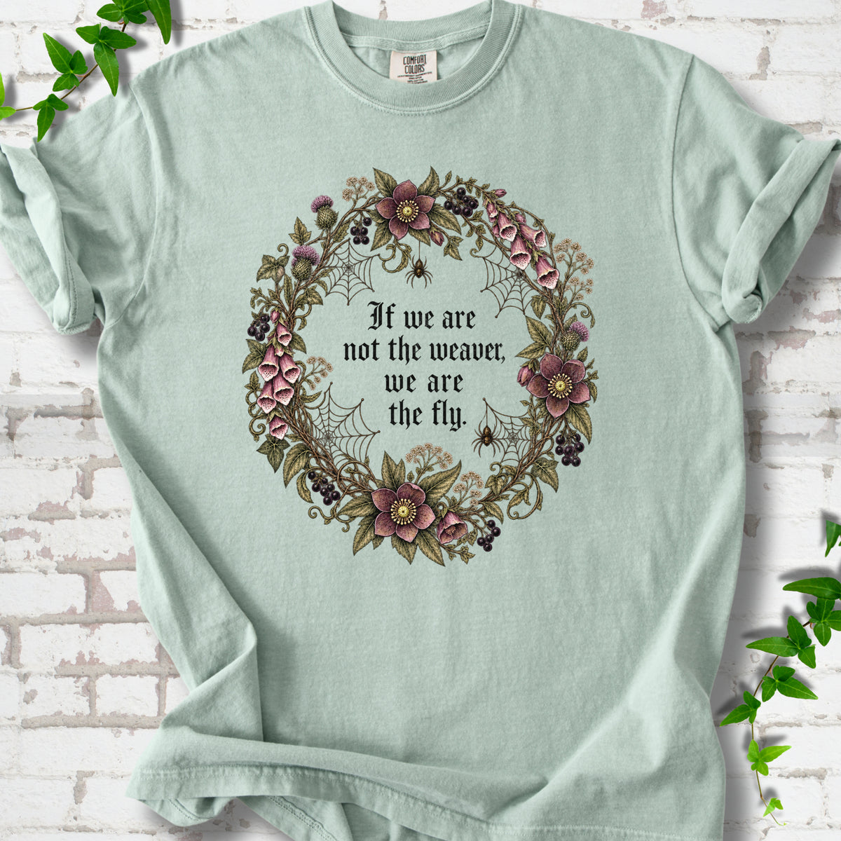 Bay T-shirt flat-lay mockup with a gothic floral wreath, spider webs and "If we are not the weaver, we are the fly" lettering.