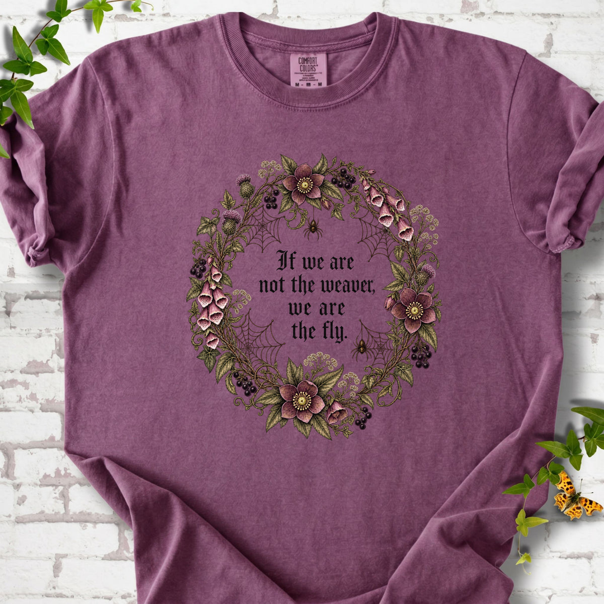 Berry T-shirt flat-lay mockup with a gothic floral wreath, spider webs and "If we are not the weaver, we are the fly" lettering.