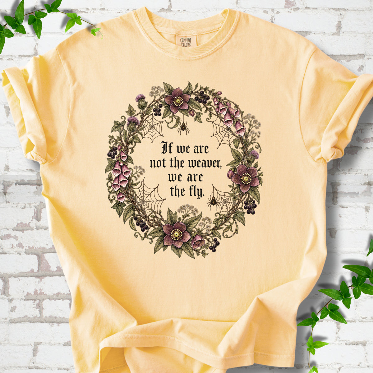 Butter T-shirt flat-lay mockup with a gothic floral wreath, spider webs and "If we are not the weaver, we are the fly" lettering.
